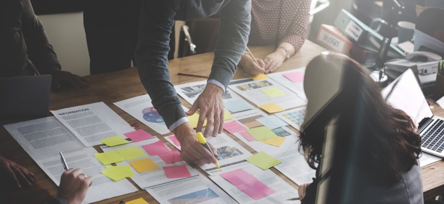 Group of business people stood around a table covered in paper and post-it notes.